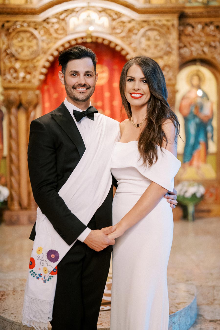 A couple in formal attire smiling inside a decorated venue with ornate wooden arches, wearing traditional embroidered ceremonial sashes.