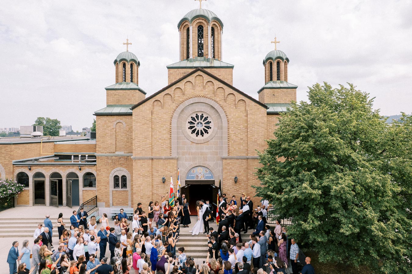 A large crowd gathers at the entrance of a historic Orthodox church for a wedding celebration. The bride and groom stand on the steps, surrounded by friends and family, with beautiful architectural details and towering spires in the background.