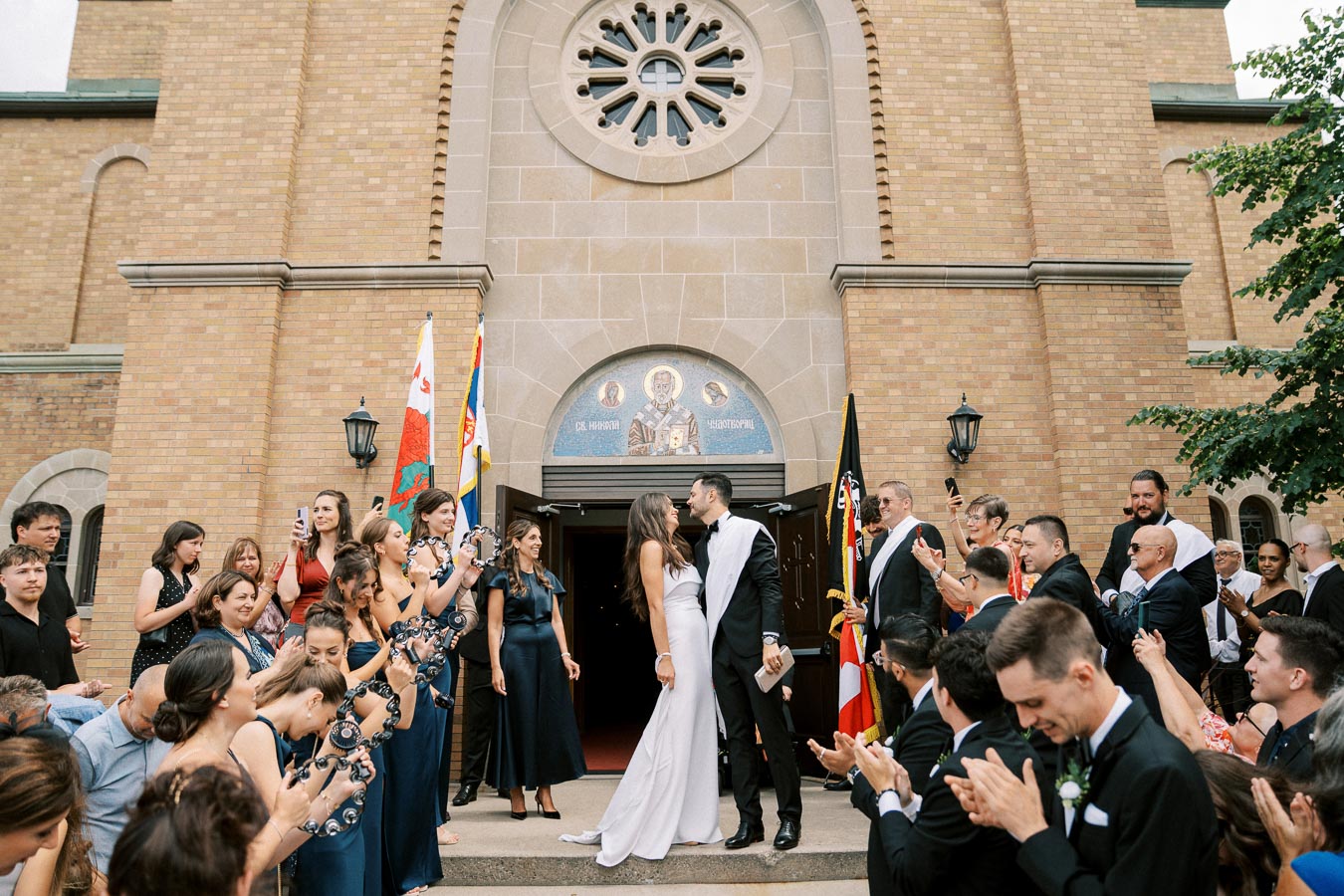 A newlywed couple kisses in front of a brick church as an excited crowd of friends and family applauds and takes photos, capturing the joyful wedding celebration.