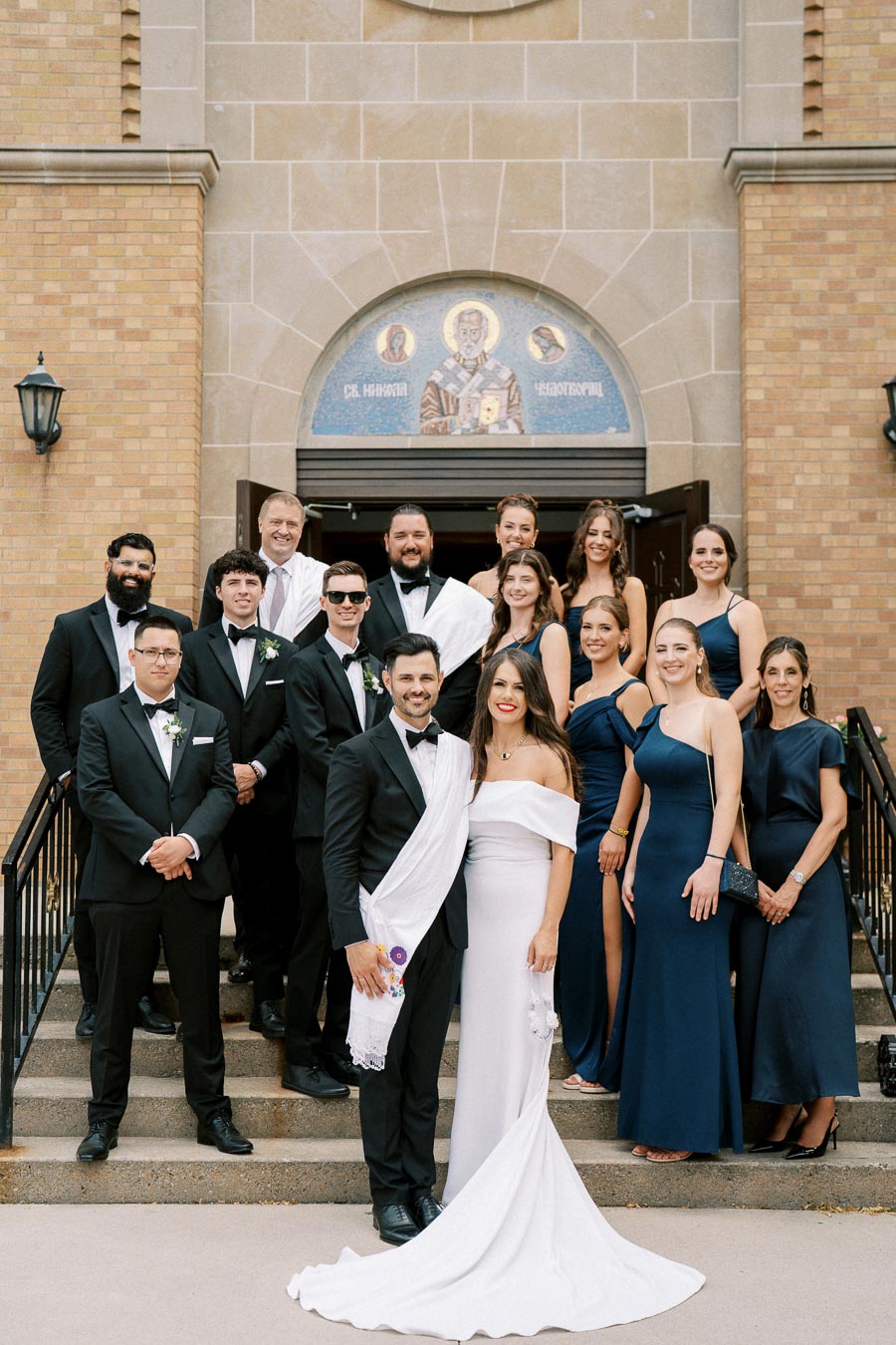 A wedding party posing in front of a church entrance, featuring the groom in a black tuxedo and the bride in an elegant white gown, surrounded by bridesmaids in navy dresses and groomsmen in black suits.