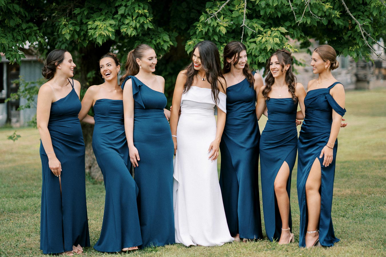 A bride in a white gown stands with her bridesmaids in matching blue dresses, smiling and laughing under a lush green tree during a wedding photo shoot outdoors.
