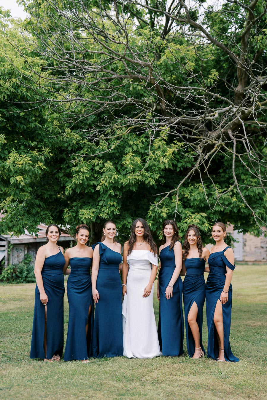 A group of seven women in elegant formal dresses, with six wearing matching navy dresses and one in a white gown, pose under a lush green tree outdoors, showcasing a coordinated bridal party theme.