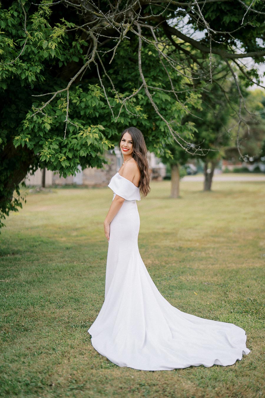 A bride in a flowing white dress smiles while standing under a lush green tree in a serene outdoor setting.