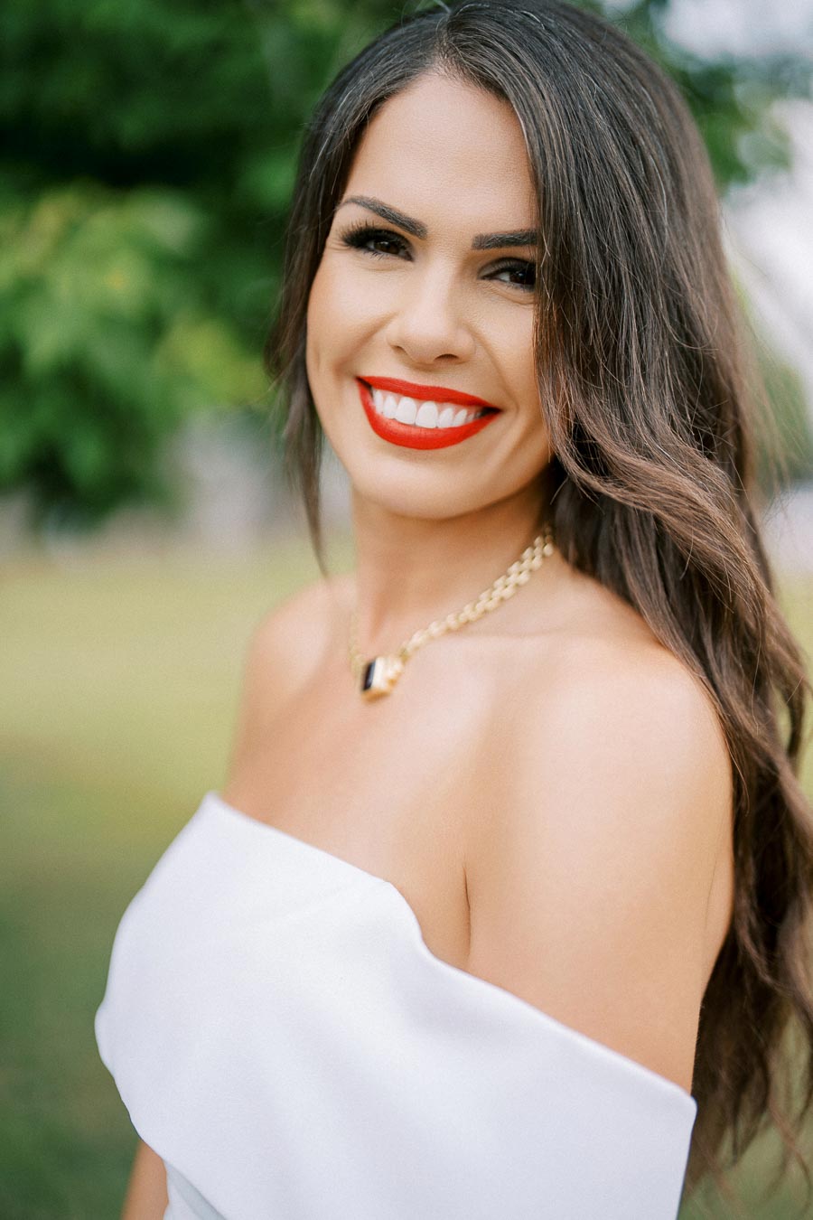 A woman with long brown hair and bright red lipstick, wearing a white off-the-shoulder top and a gold necklace, smiles warmly at the camera against a blurred outdoor background.