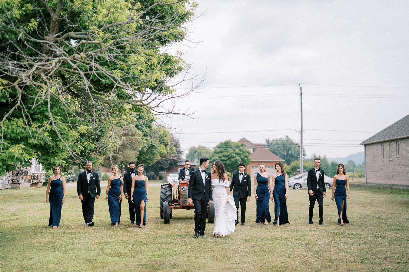 A wedding party walking on a grassy field, featuring bridesmaids in blue dresses and groomsmen in black suits. In the center, the bride and groom are holding hands, with a rustic tractor in the background, set against a backdrop of lush greenery and a cloudy sky.