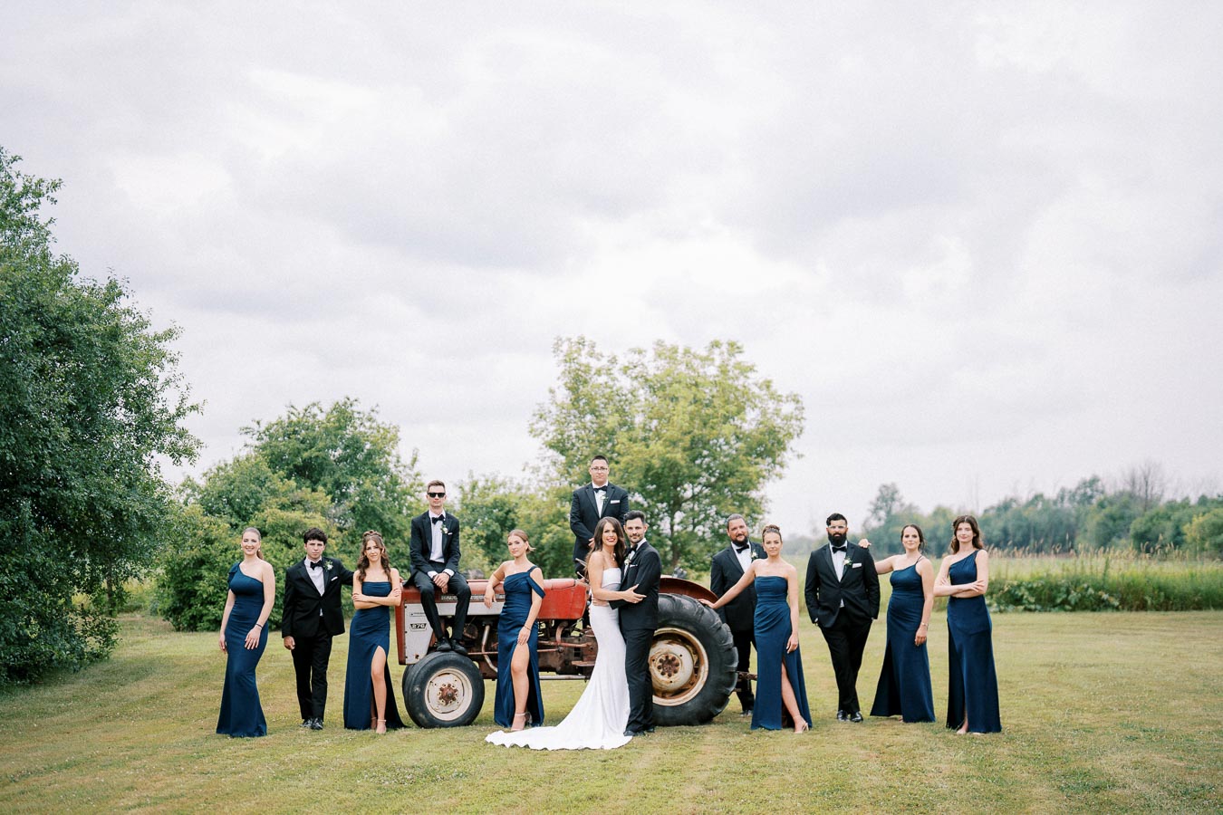 Wedding party posing outdoors with a tractor, featuring the bride in a white dress, groom in a suit, and bridesmaids in blue dresses, all set against a lush green landscape.
