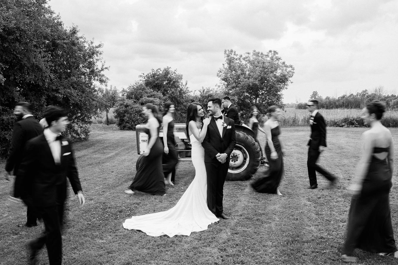 Black and white photo of a smiling bride and groom embracing in a garden setting, surrounded by moving wedding party members in elegant attire, with a vintage tractor in the background.