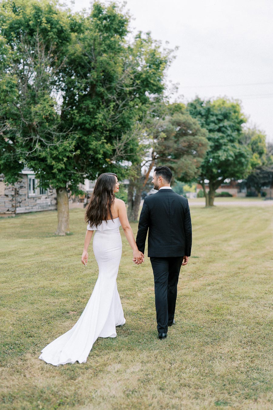 Couple holding hands while walking on grass, with the woman in a white dress and the man in a black suit, surrounded by lush green trees.