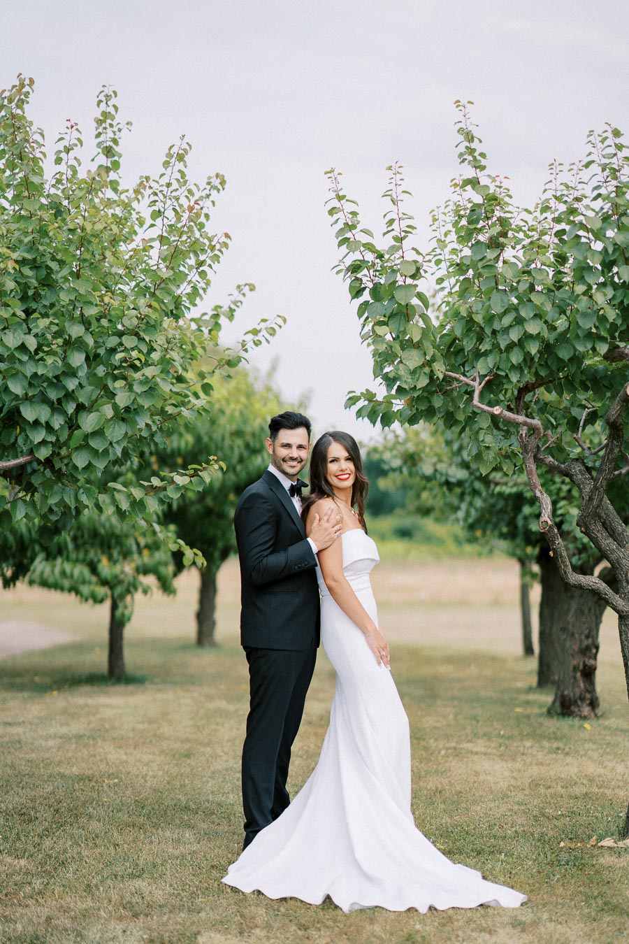 A bride and groom smiling and embracing in an outdoor garden setting, with the bride in a white gown and the groom in a black suit, surrounded by lush green trees under a clear sky.