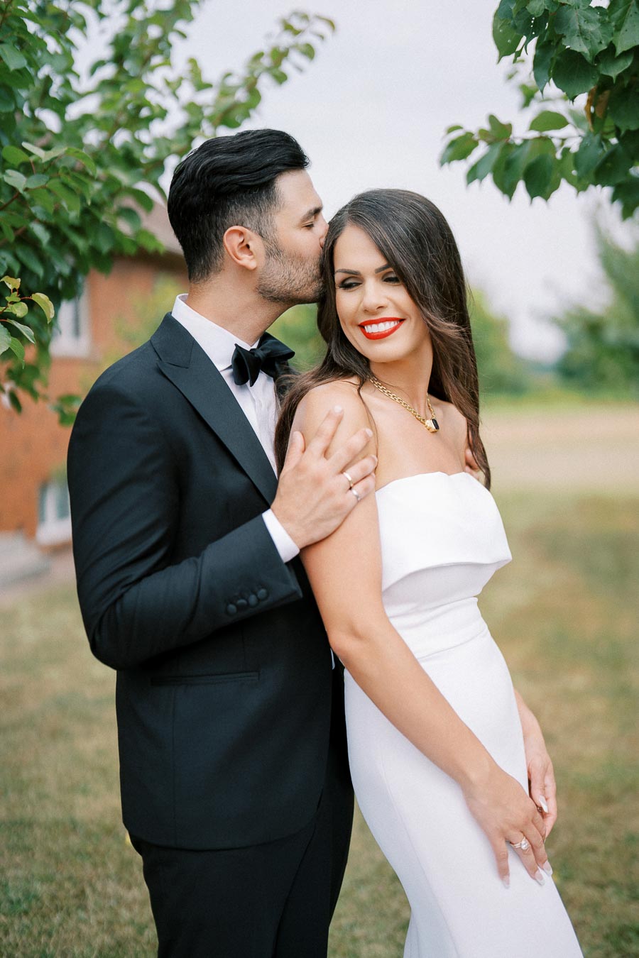 Elegant couple posing outdoors, man in black tuxedo gently kissing woman's forehead, woman in white dress smiling brightly, surrounded by green foliage.