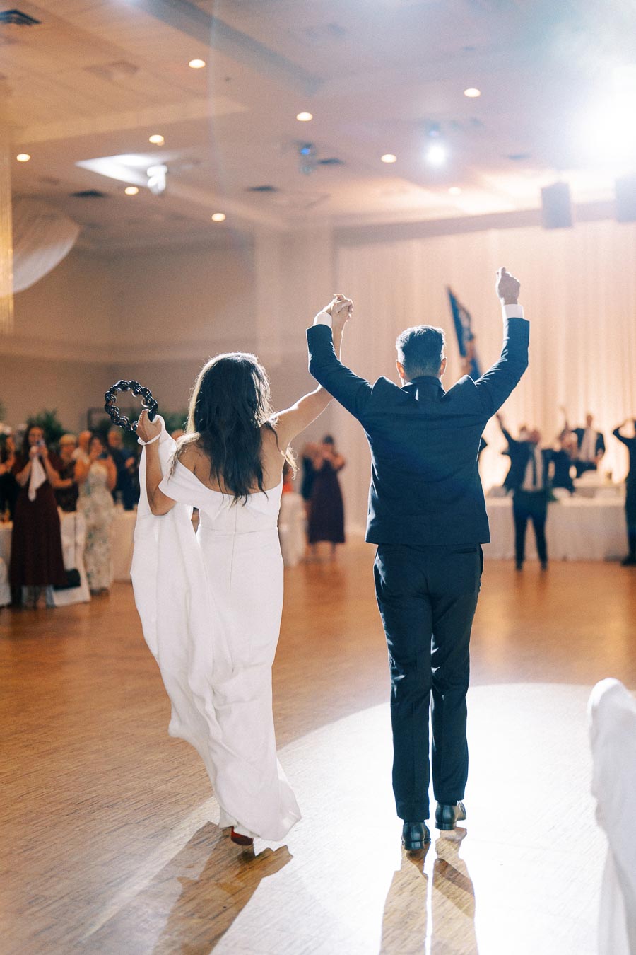 A newlywed couple celebrates by holding hands and raising their arms together on the dance floor during a wedding reception, captured with bright lighting and guests clapping in the background.