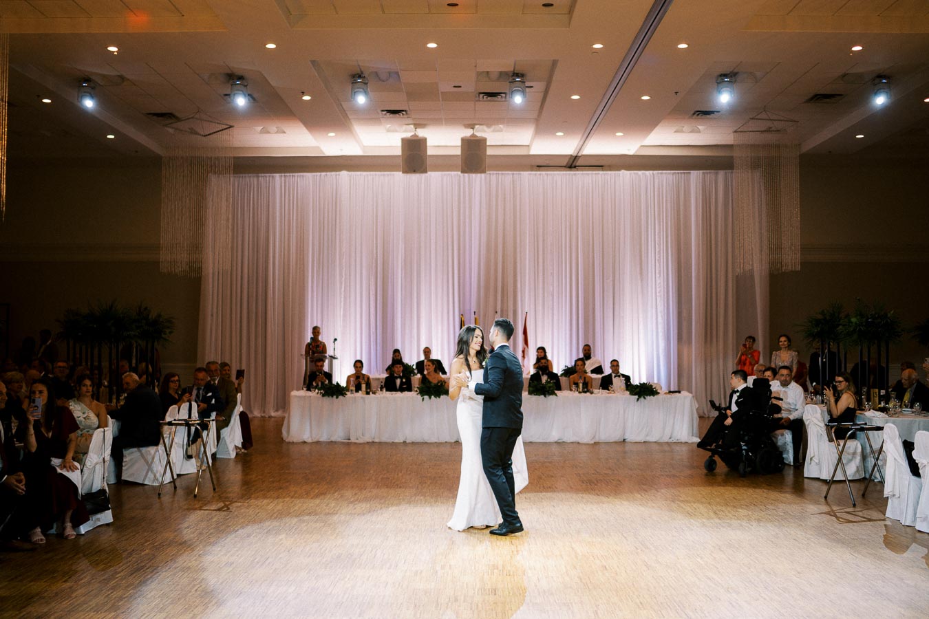 A bride and groom share their first dance at a wedding reception in an elegant ballroom, surrounded by seated guests and a decorated head table with a white drapery backdrop.