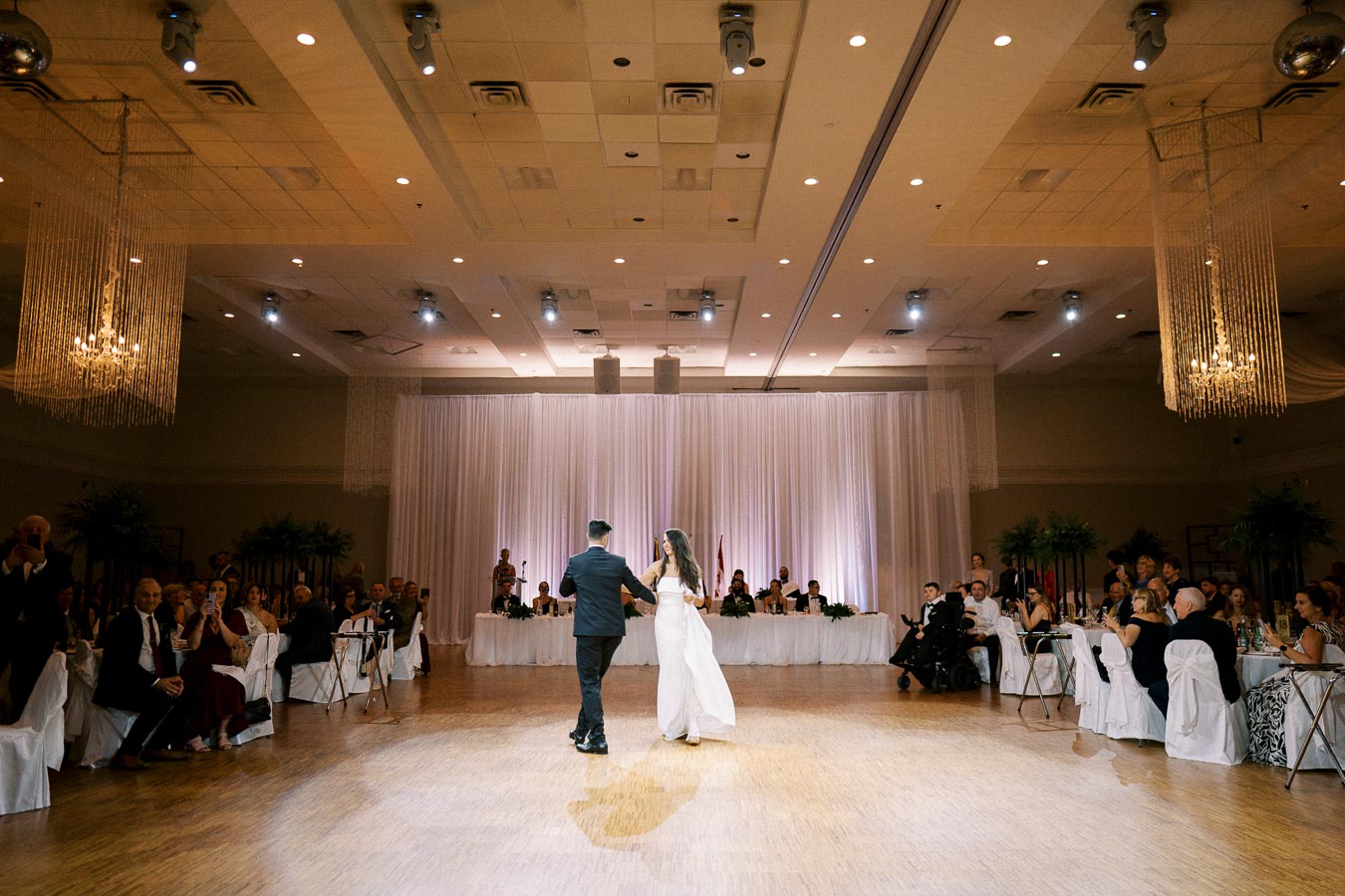 A couple performs a romantic first dance at a wedding reception in an elegantly decorated ballroom, surrounded by seated guests and soft, ambient lighting.