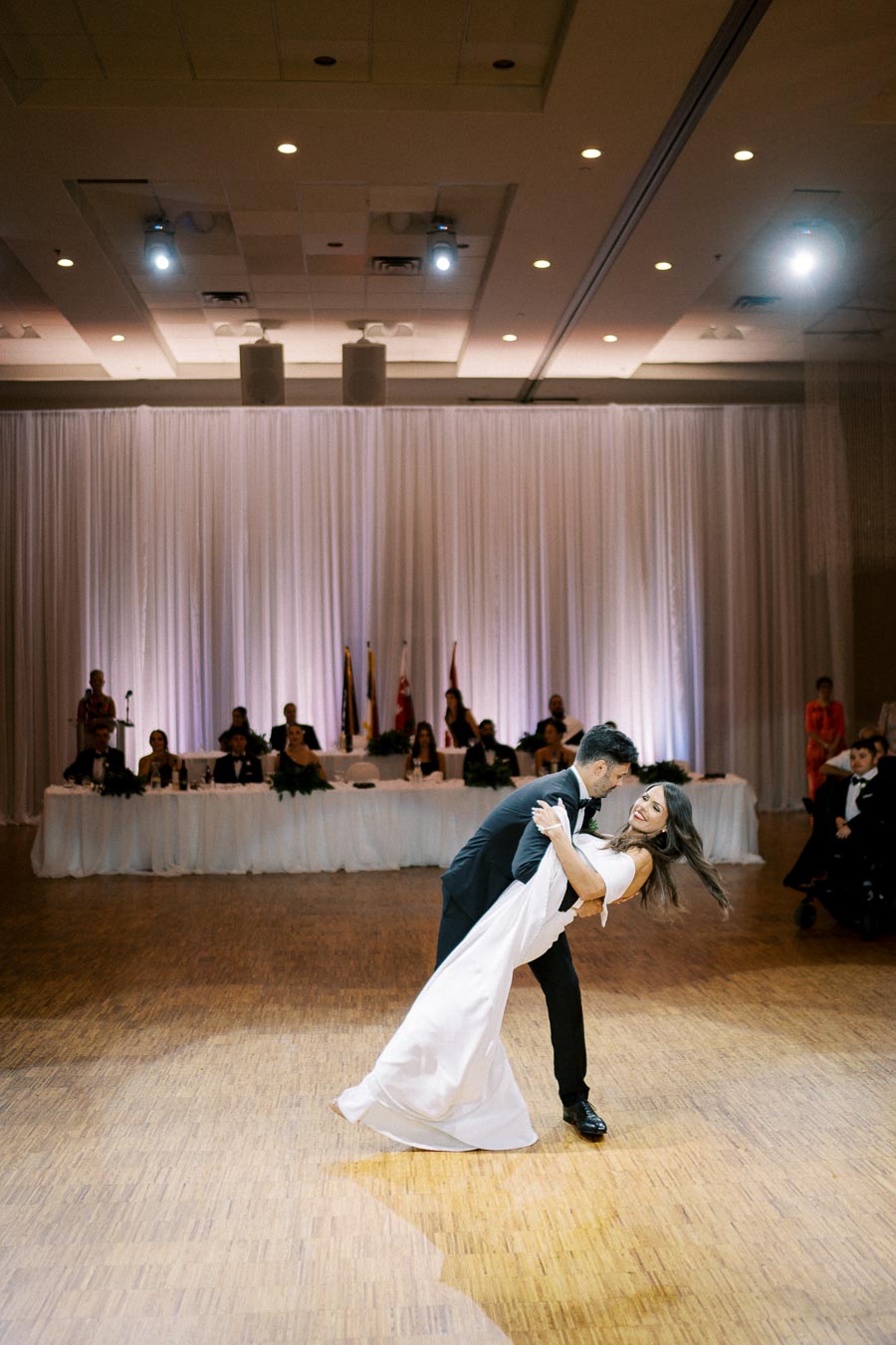 A bride and groom share a romantic dance at their wedding reception, gracefully dipping on a polished wooden floor with a backdrop of elegant white drapes and seated guests.