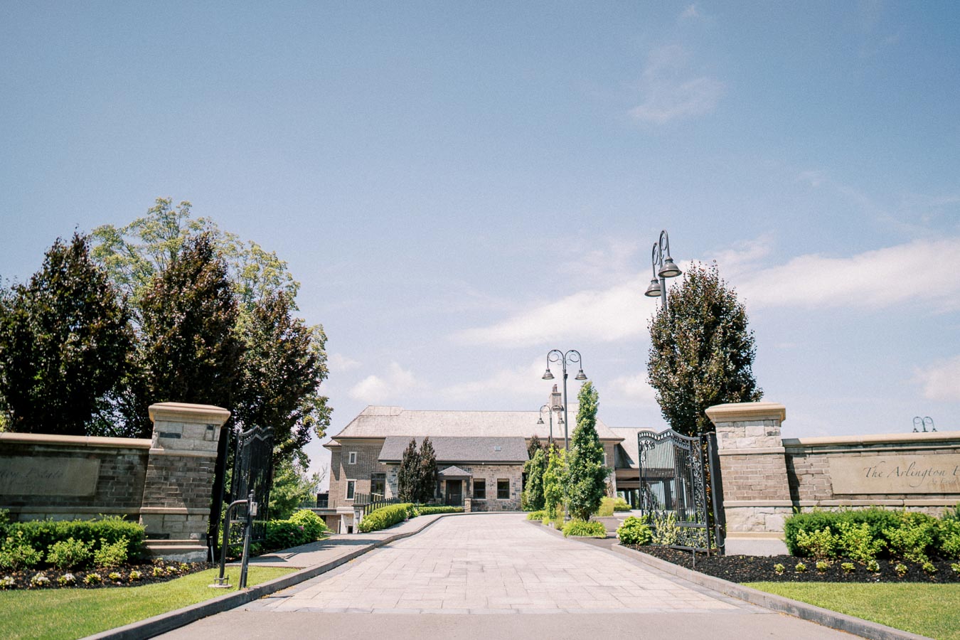 Grand entrance to The Arlington Estate with paved driveway, surrounded by manicured gardens and tall lamp posts under a clear blue sky.