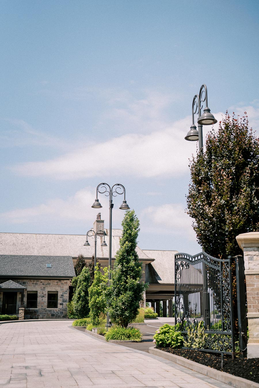 Elegant stone building with decorative black iron gate and vintage street lamps under a clear blue sky, surrounded by manicured greenery and a paved driveway.