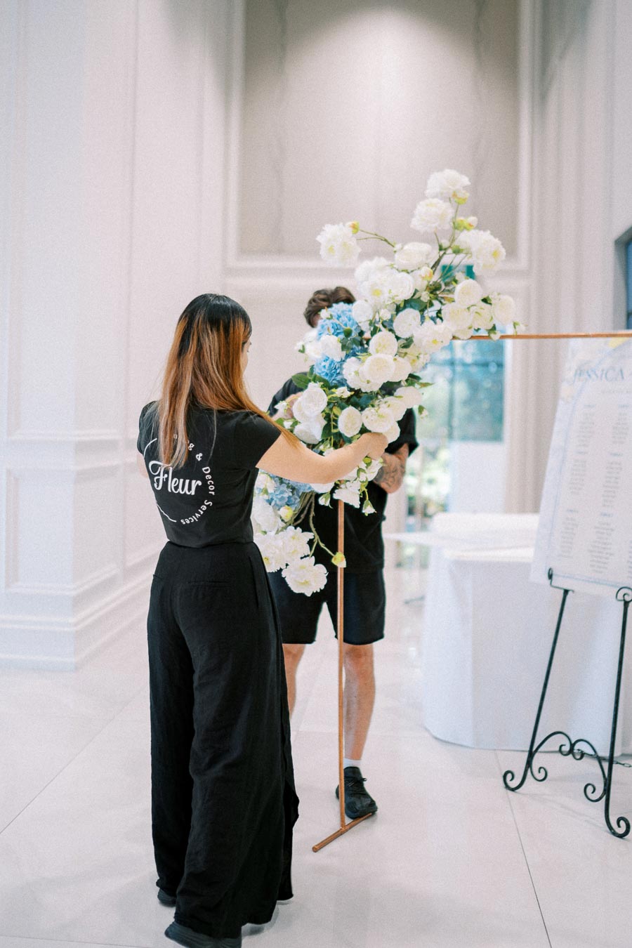 A florist arranging a large bouquet of white and blue flowers in an elegant indoor setting, wearing a black outfit with Fleur branding, emphasizing professional event decor services.