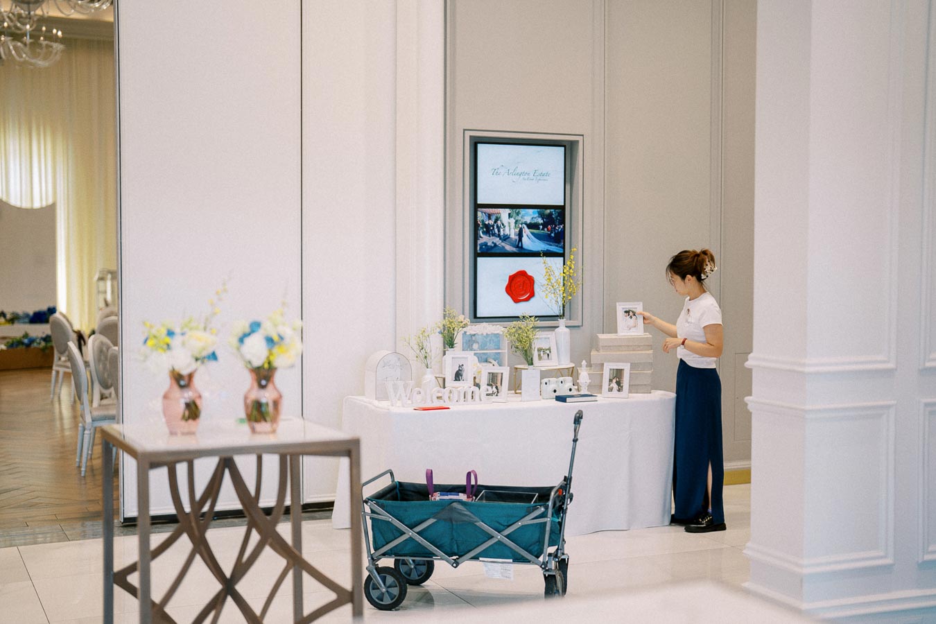 Elegant event setup with a table displaying decorative photo frames and flowers, a woman organizing items, and a wagon nearby in a well-lit room.