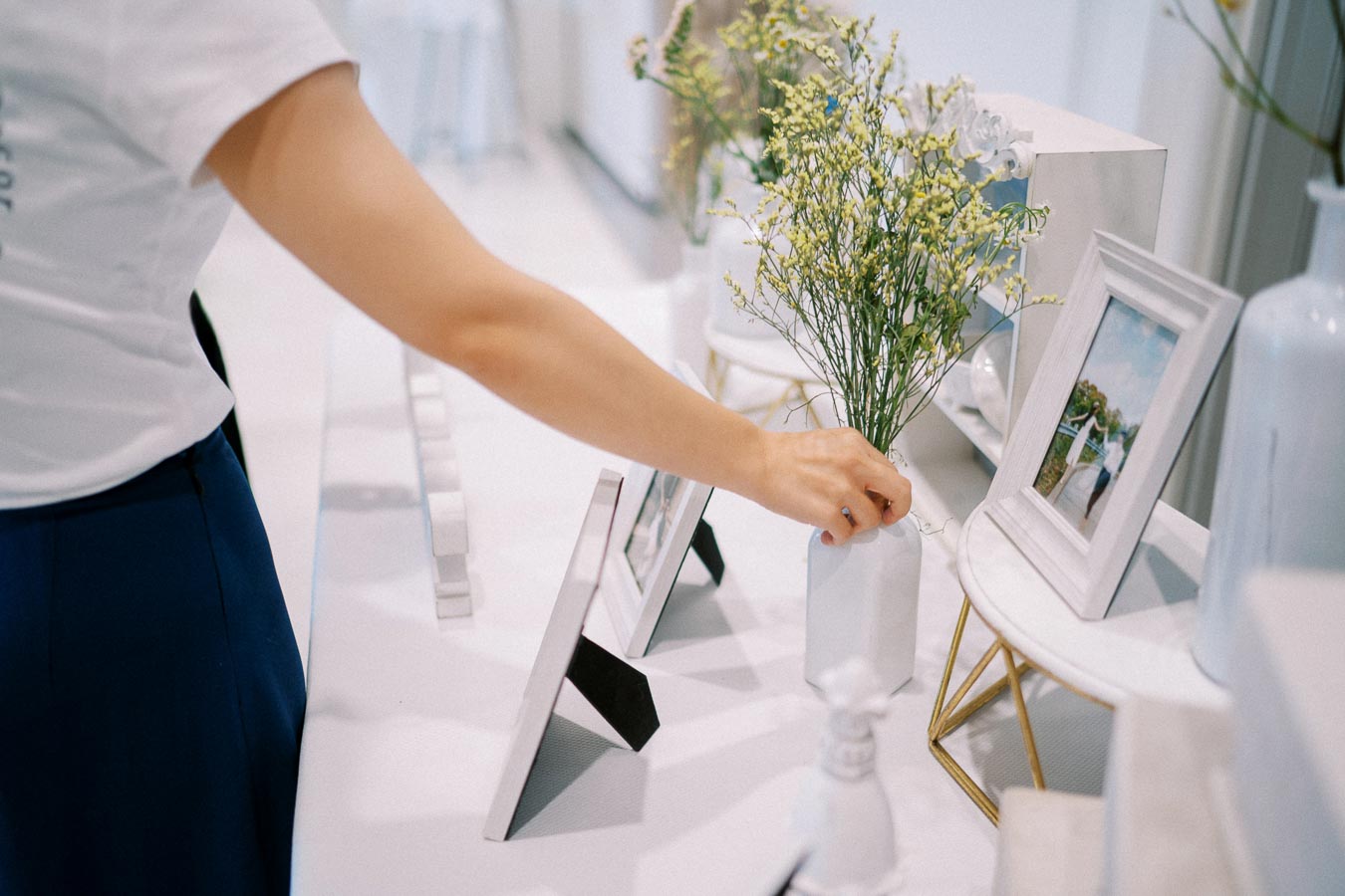 A person arranging flowers in a white vase on a table decorated with picture frames and minimalist decor.