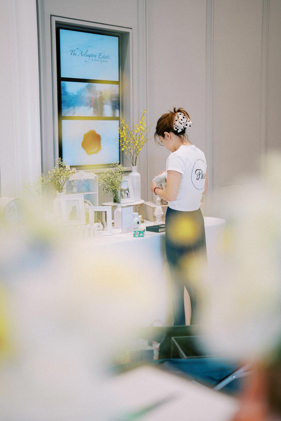 A woman in a white shirt arranges decorative items and flowers on a table, with a digital display showing the text “The Arlington Estate” in the background, interior decor setup.