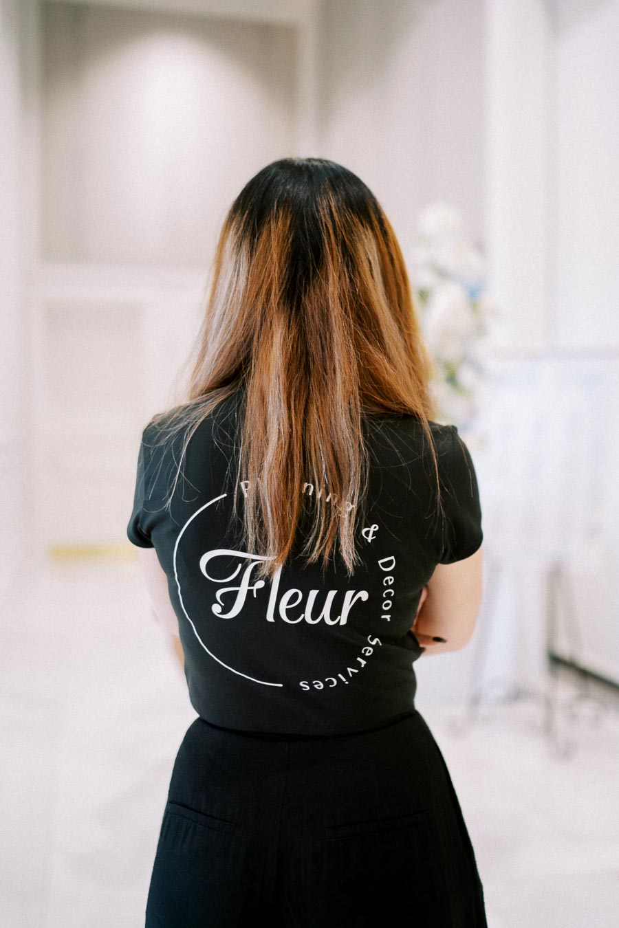 A woman with long hair wearing a black shirt with Fleur printed on the back, standing indoors in a well-lit space.