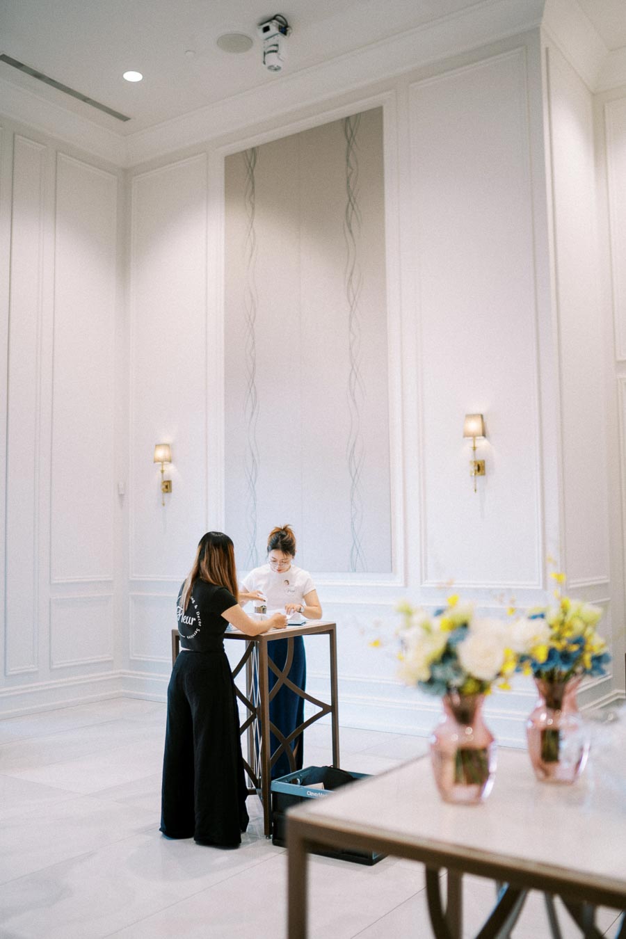 Two women working together at a high table in a bright, elegant room with floral decorations.