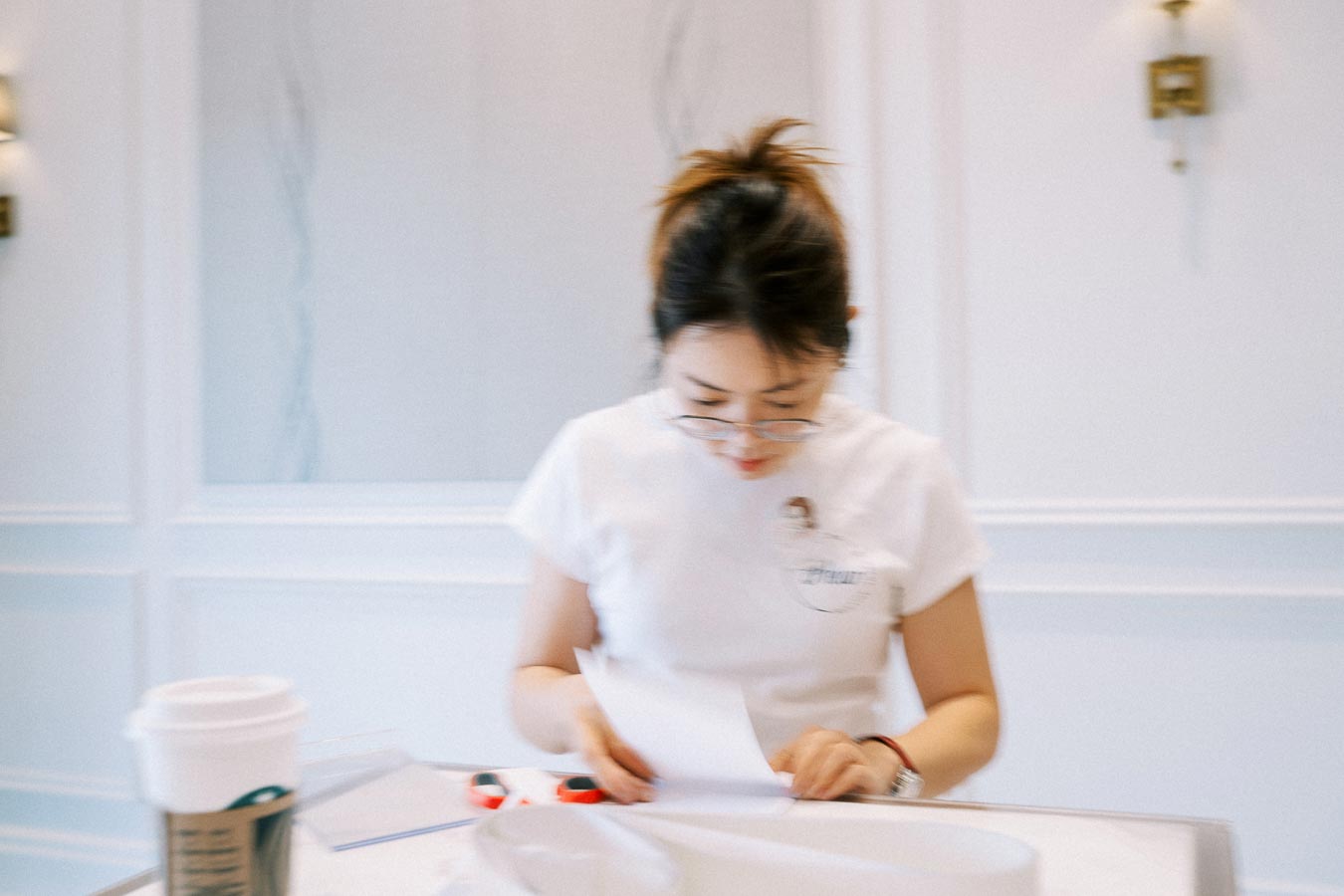 Blurred image of a woman in glasses focusing on papers, with a cup of coffee on a table in a bright room with white walls.