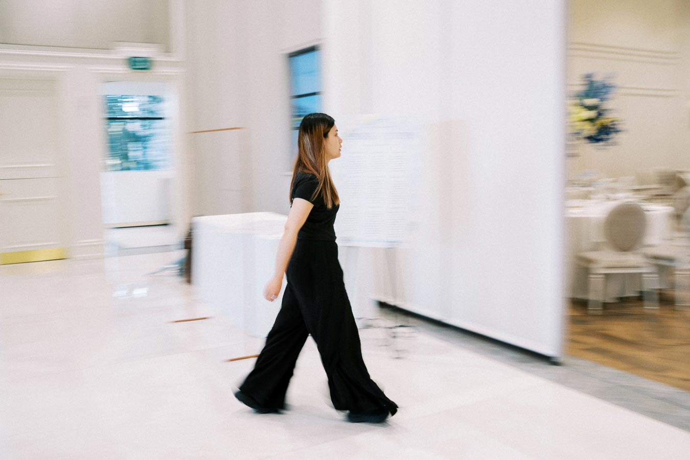 A woman in motion walks through a modern, elegant indoor space with white walls and large windows, dressed in a black outfit, capturing a sense of speed and purpose in a softly blurred background.
