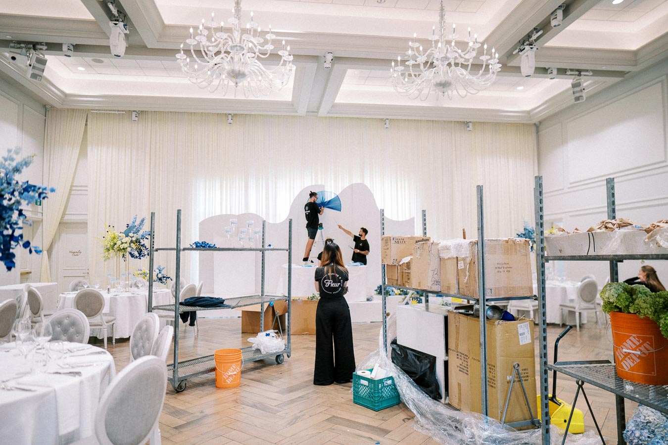 Elegant event setup in progress in a sophisticated venue with chandeliers and draped curtains, featuring a team preparing decorations and arranging tables with floral centerpieces and seating.