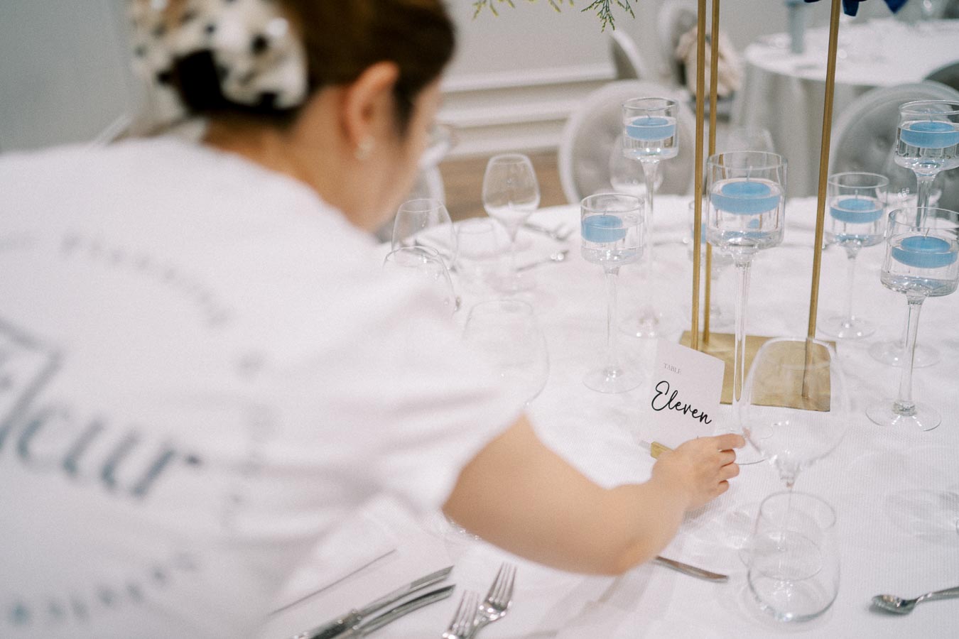 A person arranging a card labeled Table Eleven on a well-set dining table featuring elegant glassware and floating blue candles, in a sophisticated event setting.