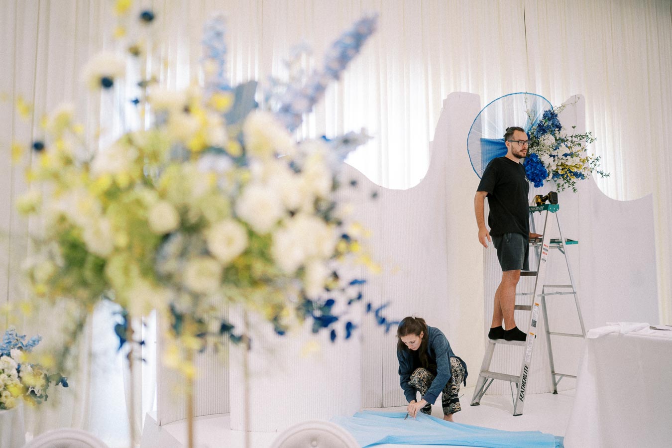 A man stands on a ladder adjusting floral decorations with yellow and blue flowers, while a woman kneels on the floor arranging blue fabric in a decorated room with white drapery.