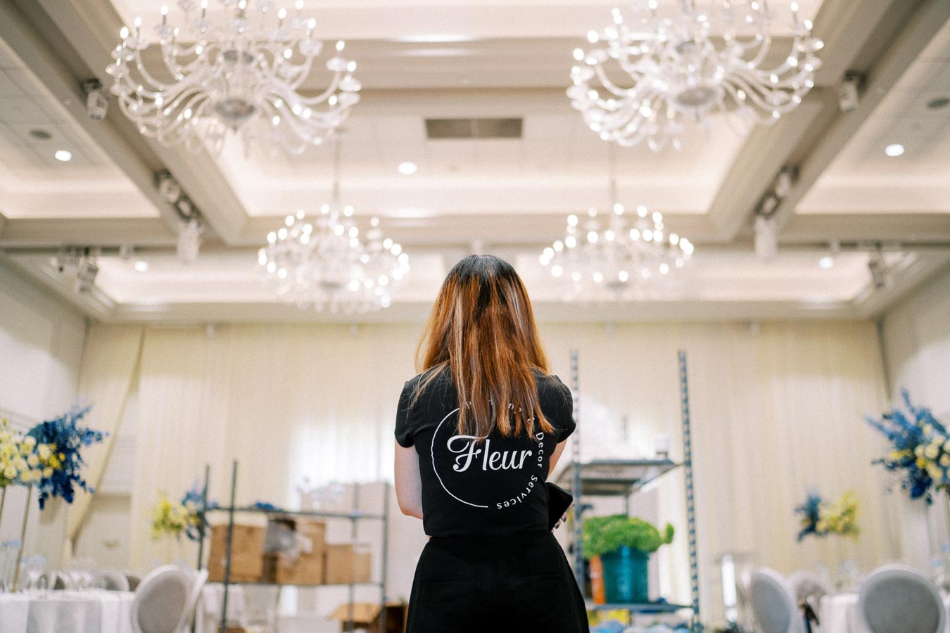 Rear view of a woman in a black shirt with a logo, standing in an elegantly decorated room with chandeliers and floral arrangements.