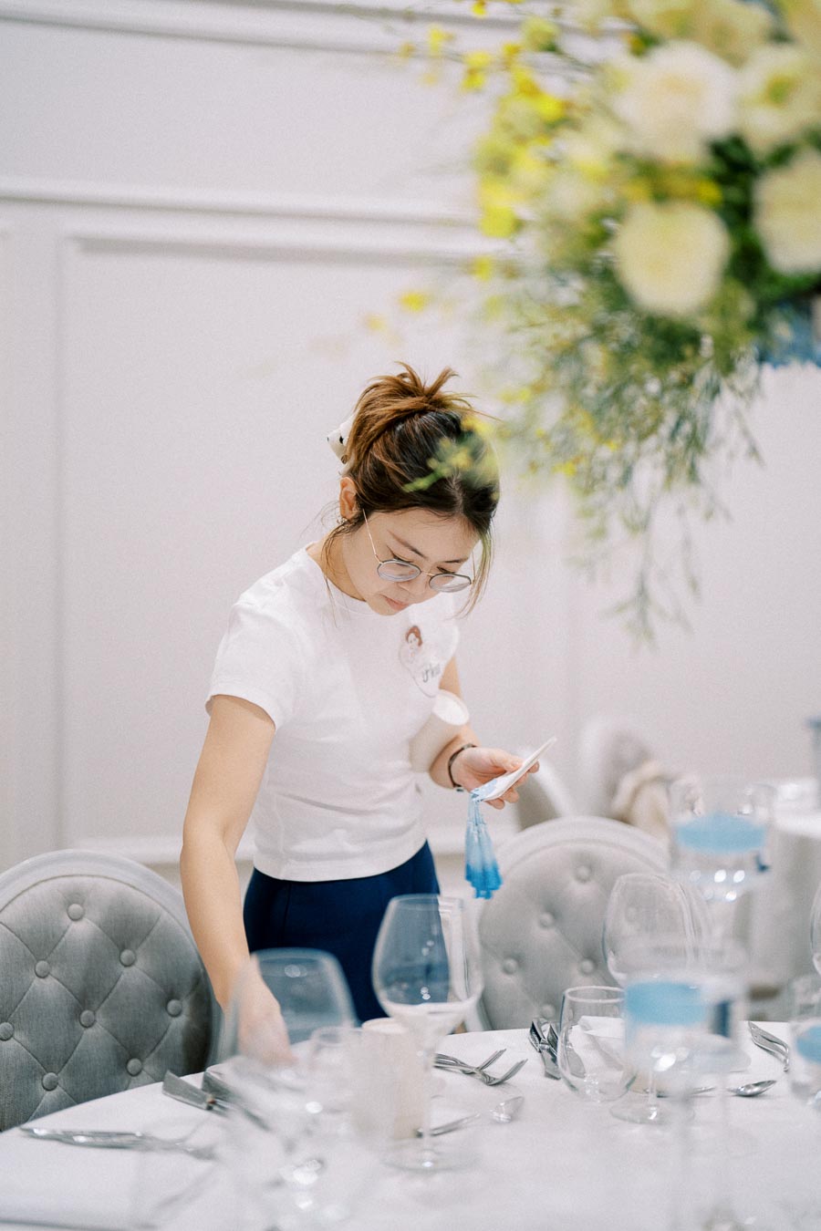 Young woman setting a sophisticated dining table with white linen, glassware, and utensils, in an elegantly decorated room with floral arrangements.