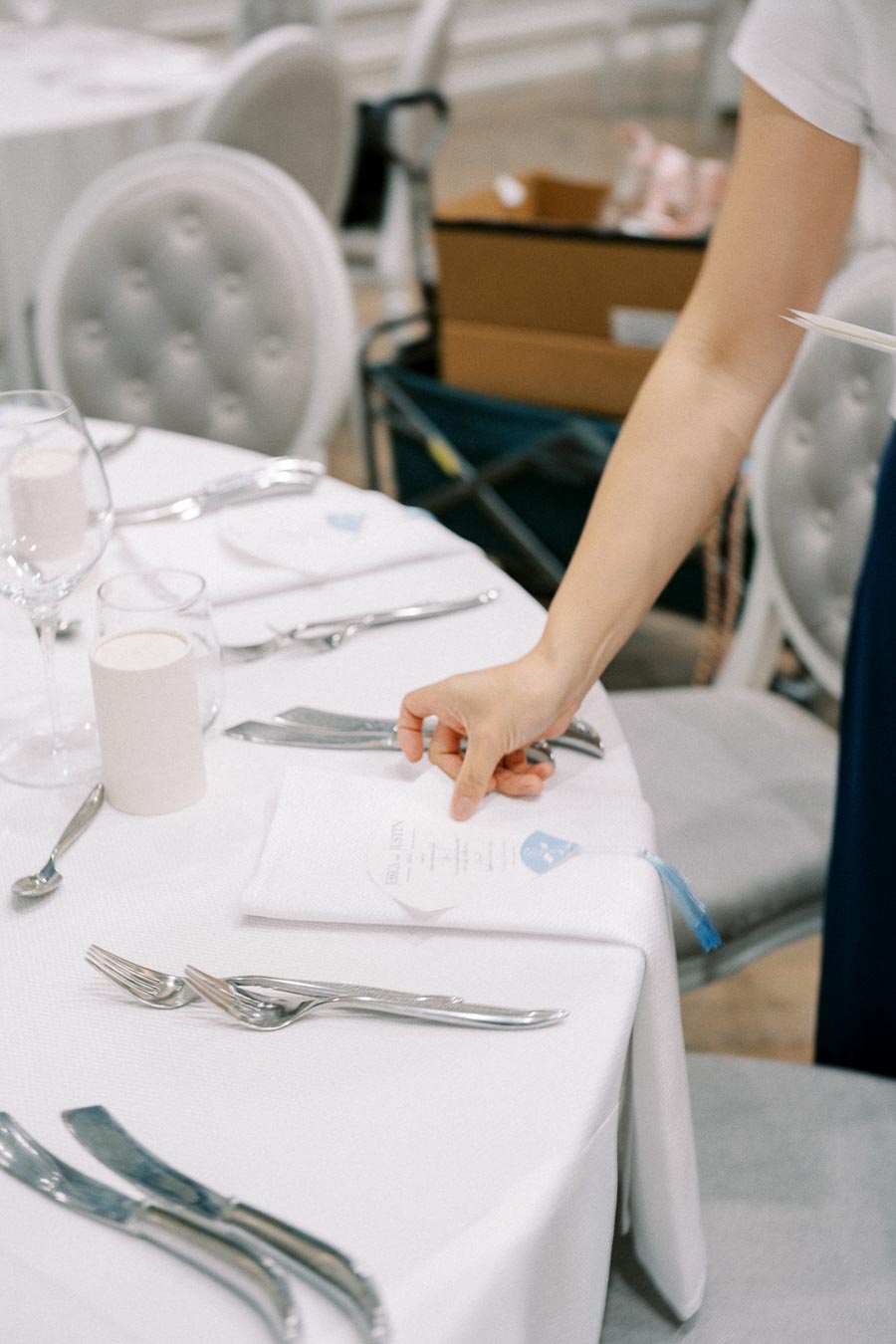 A person arranging silverware and a menu card on a neatly set white table for a formal event, with elegant chairs in the background.
