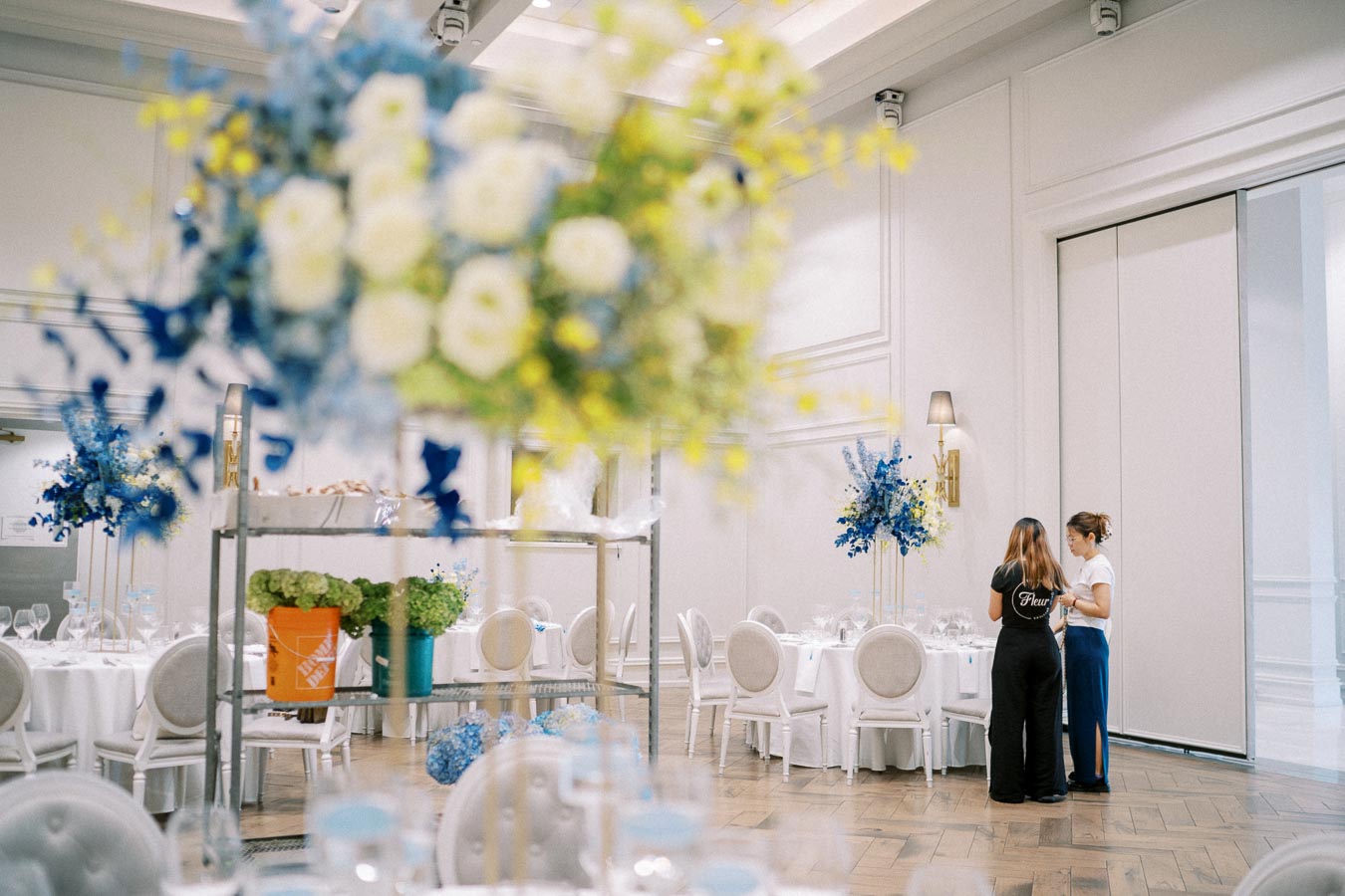 Stylish banquet hall with elegant white and blue floral arrangements, round tables set for an event, and two people discussing details in the background.