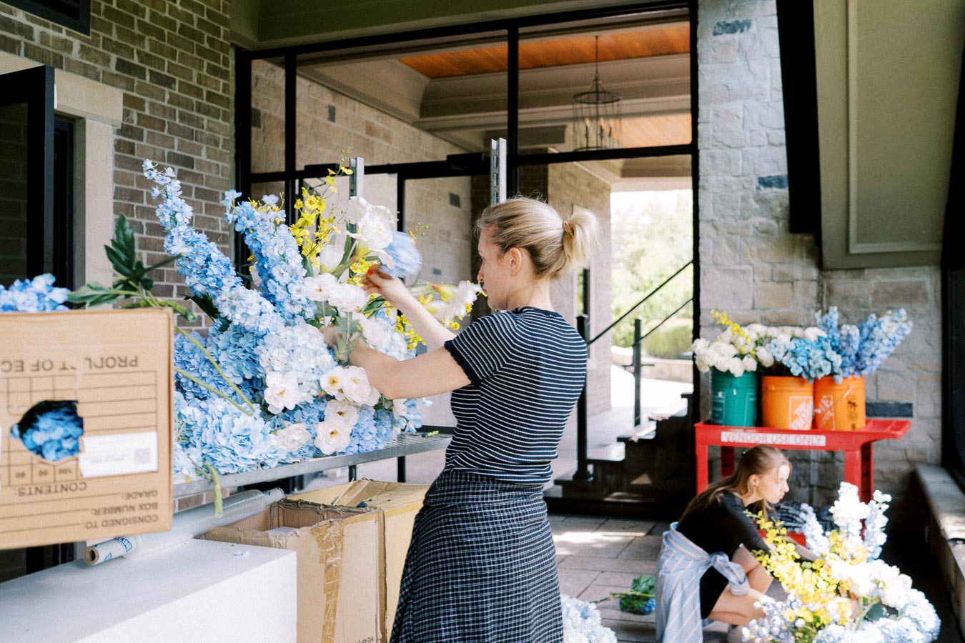Two women arranging vibrant blue and yellow flowers in a sunlit workspace with brick walls.
