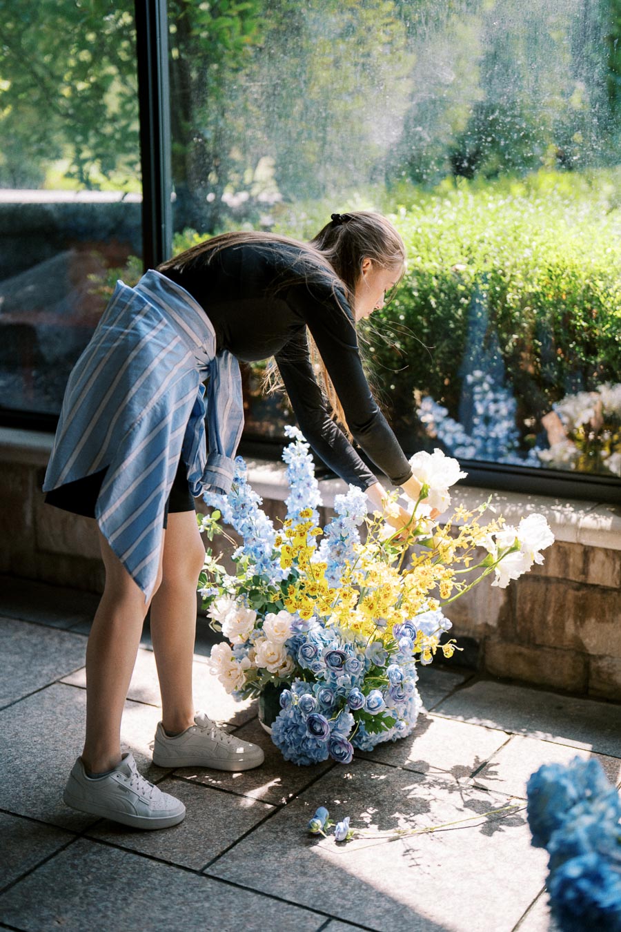 Young woman arranging a vibrant bouquet of blue, yellow, and white flowers by a sunlit window, wearing a black outfit with a blue striped shirt tied around waist.