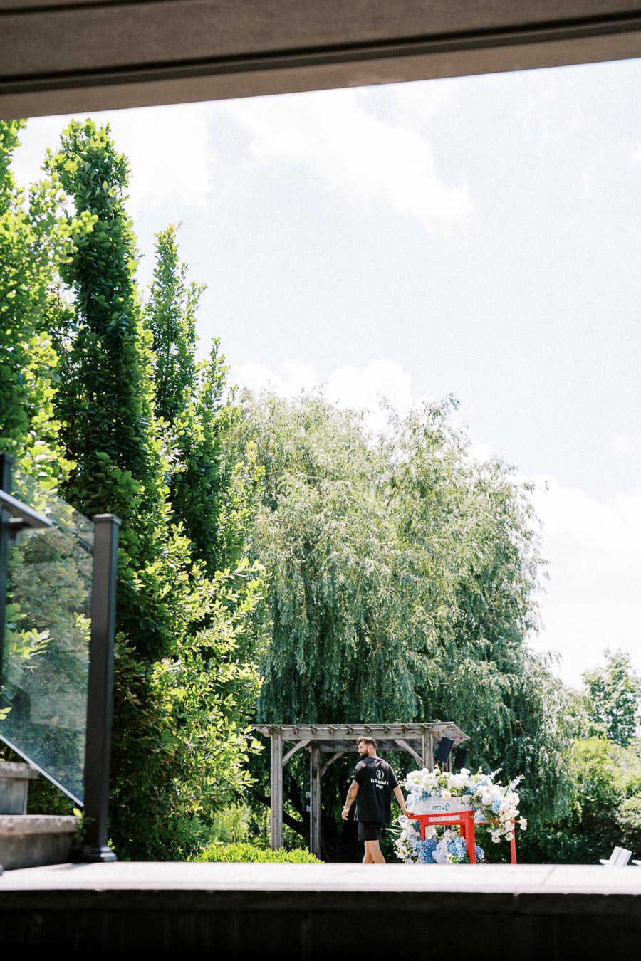 Outdoor garden scene with lush green trees and a person arranging flowers on a decorative cart under a wooden pergola.