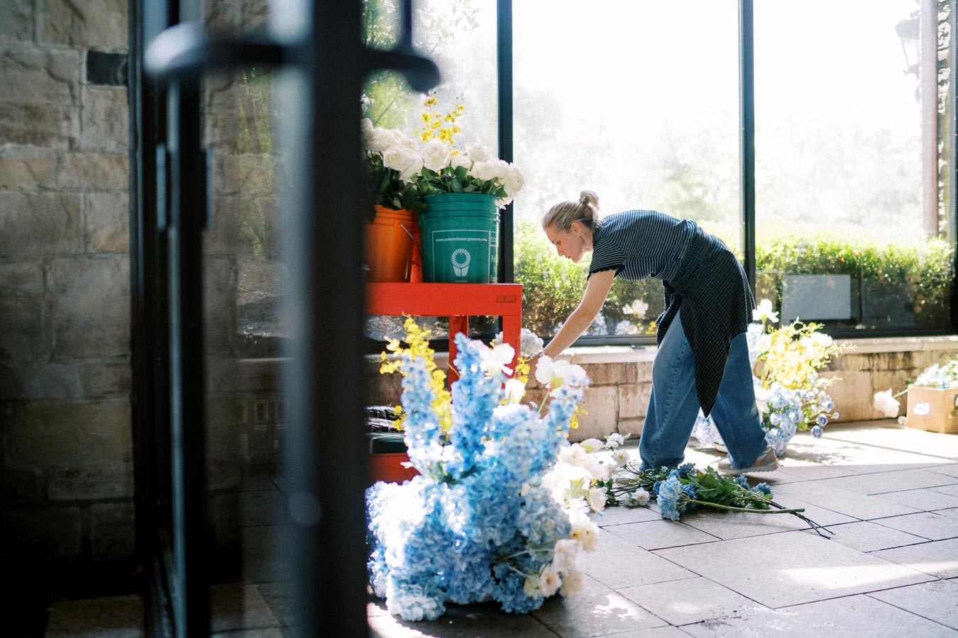 Florist arranging a variety of fresh blue, white, and yellow flowers in a sunlit greenhouse, with buckets of blooms on a red cart nearby.