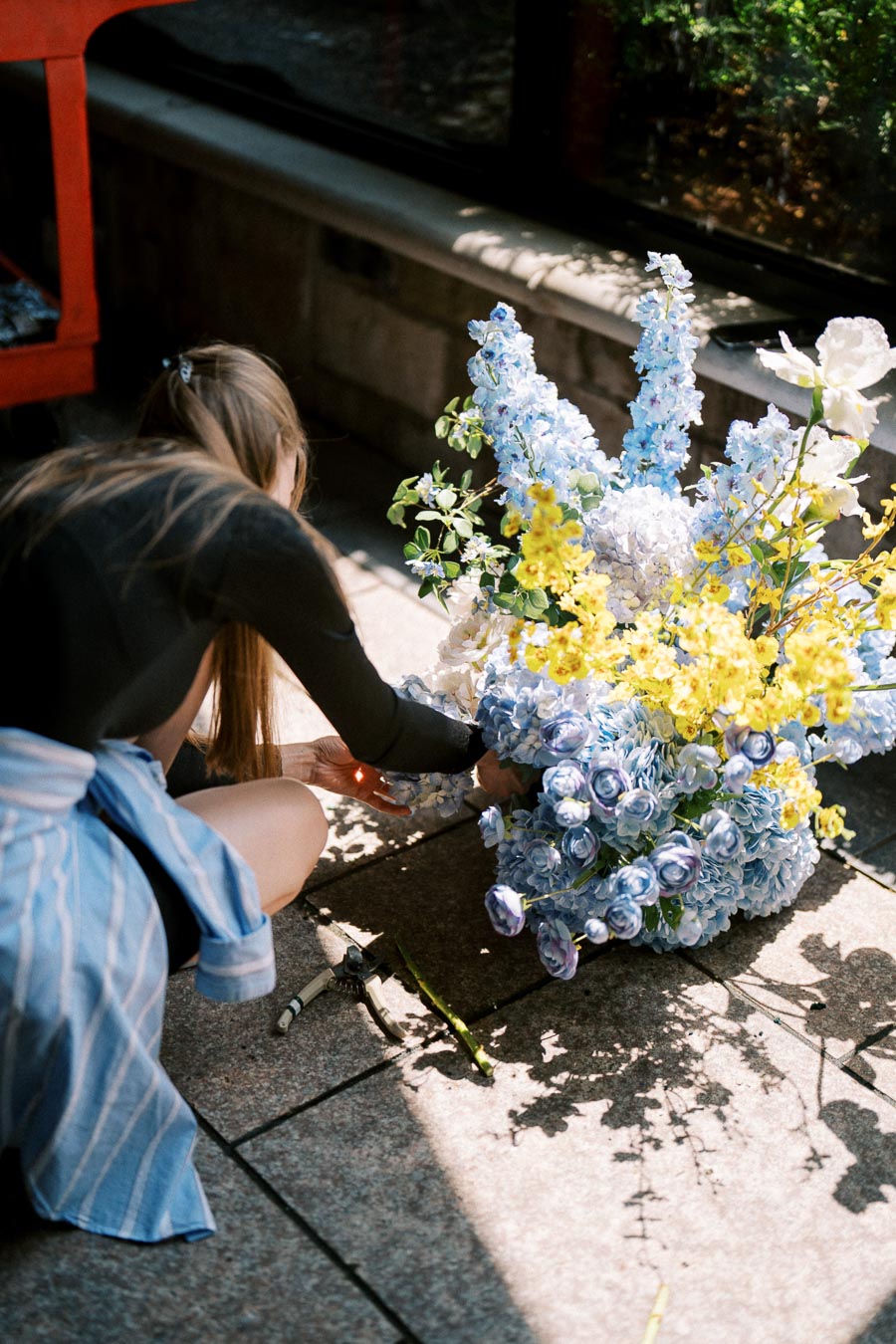 A person arranging a colorful bouquet of blue and yellow flowers on a sunlit sidewalk, with garden shears nearby.