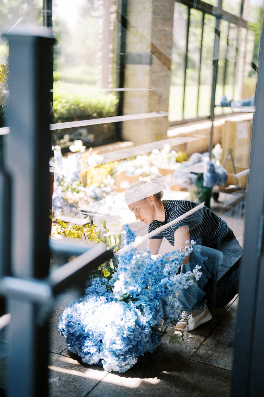 A florist arranging an elegant display of blue hydrangeas and white lilies in a bright, sunlit room with large windows.