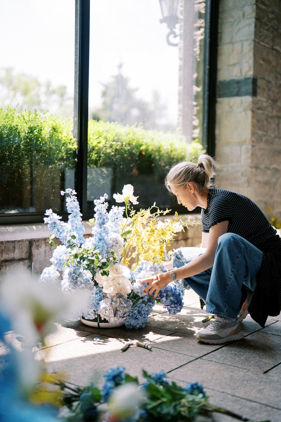Young woman arranging a vibrant floral bouquet with blue and white flowers by a sunlit window, showcasing floral design creativity.