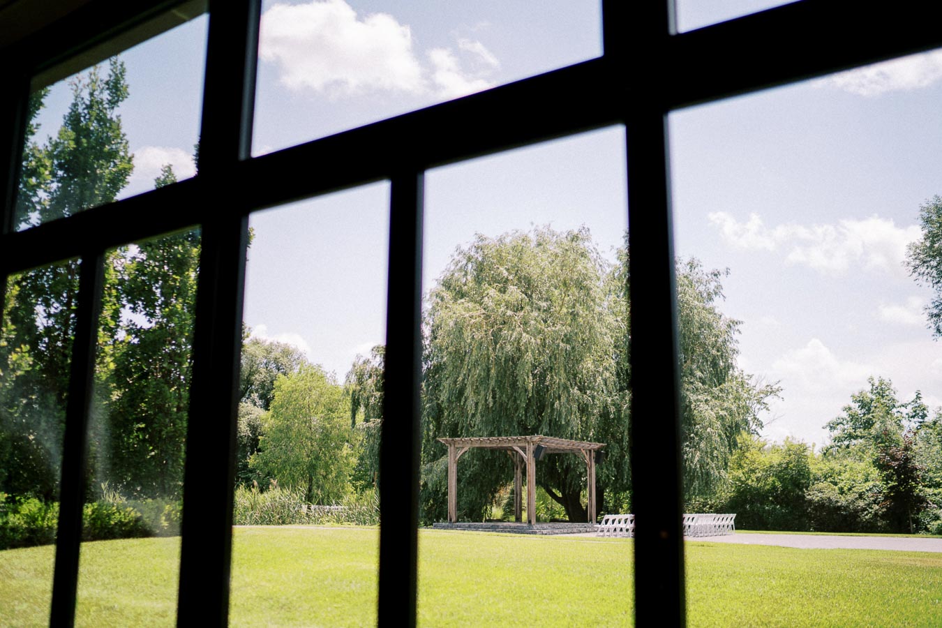 View through a window showcasing a sunny garden with a wooden pergola and lush greenery, perfect for outdoor events.