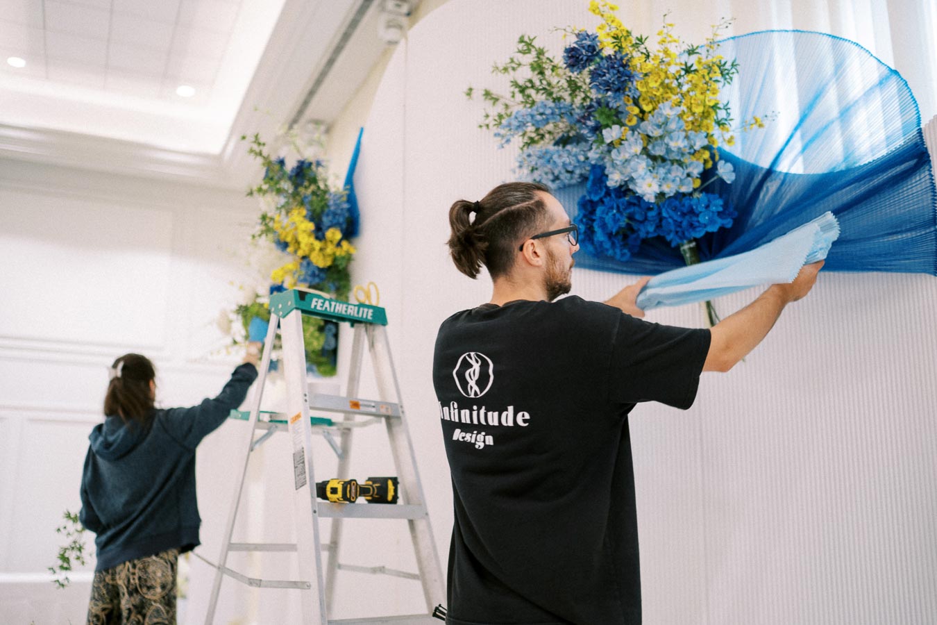 Two people setting up a blue and yellow floral arrangement for an event decoration, with one person adjusting fabric near a ladder that holds various tools in a brightly lit indoor setting.