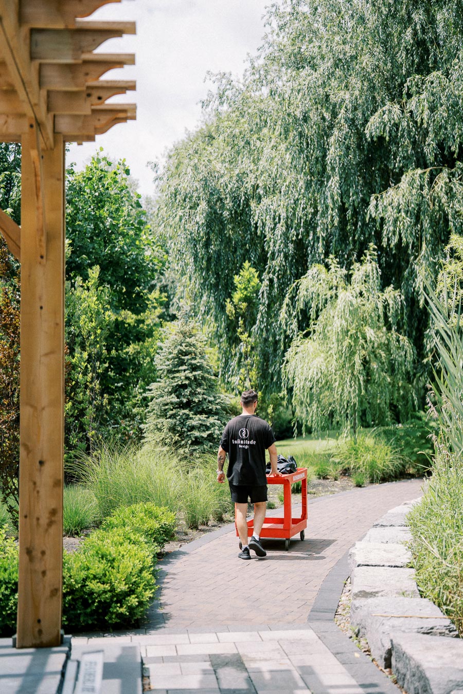 A person pushing a red cart along a paved garden pathway surrounded by lush greenery and trees under a wooden pergola.