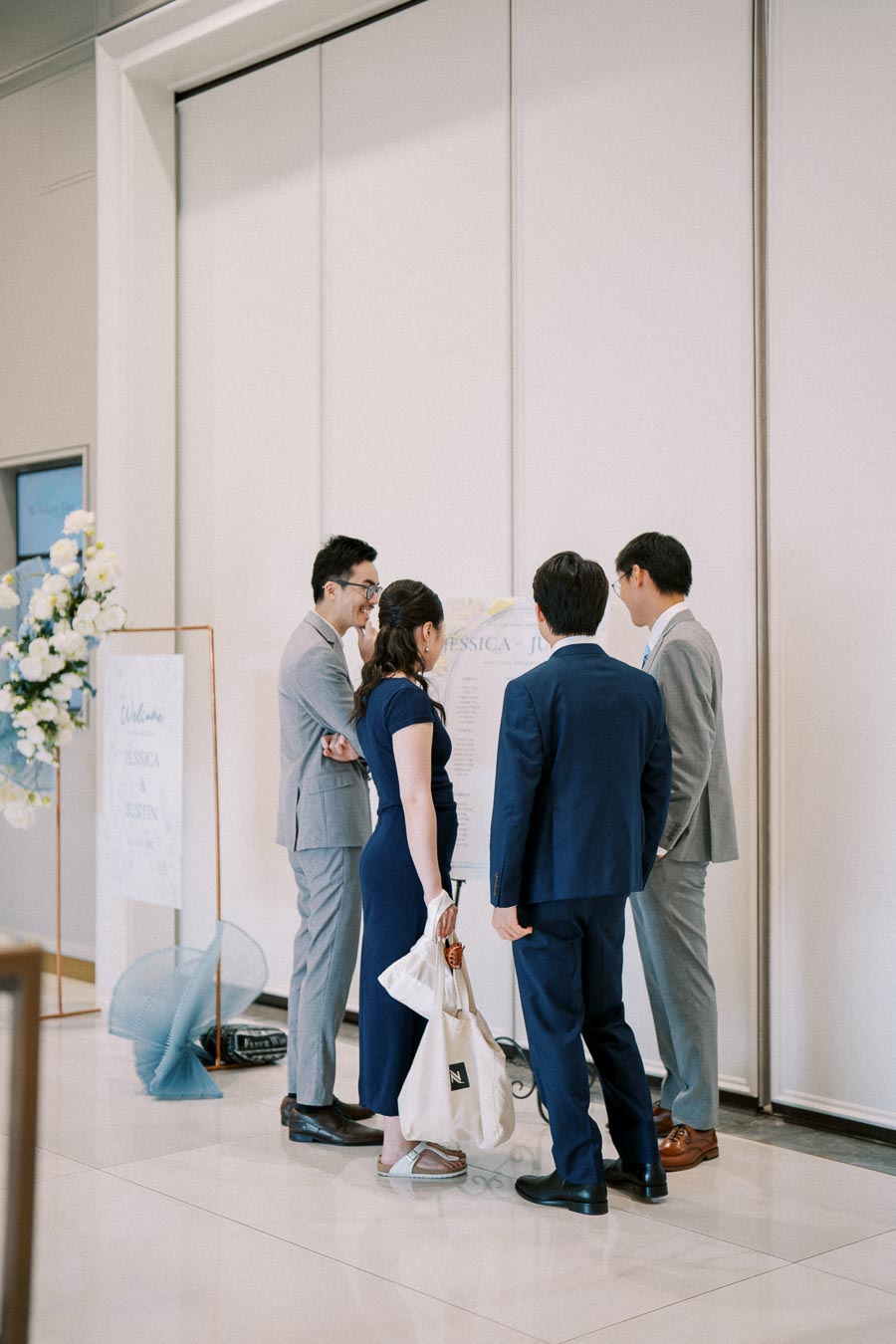 A group of four people in formal attire stand in front of a wedding seating chart at a modern indoor venue, engaged in conversation.