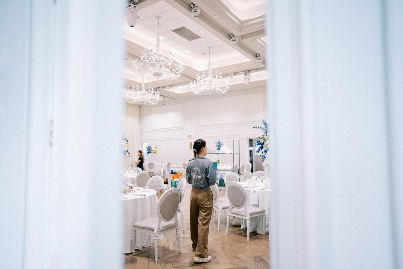 A staff member prepares a luxurious banquet hall with elegant chandeliers and white decor, featuring round tables set for an event.