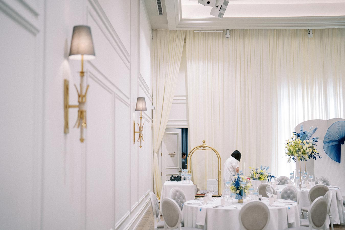 Elegant wedding reception setup with white round tables, ornate floral arrangements, and soft lighting in a luxurious venue.