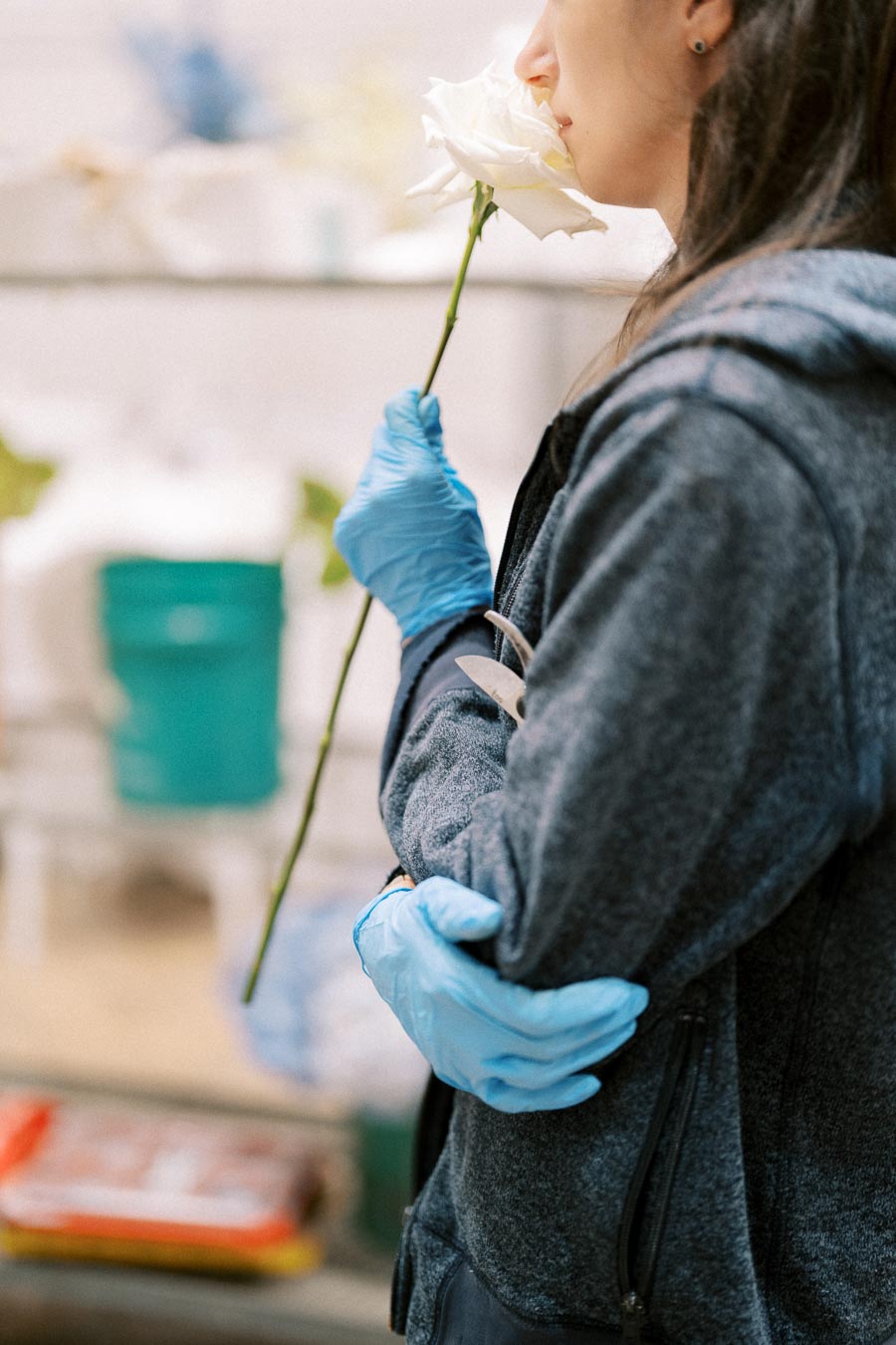 A person wearing blue gloves smelling a white rose, captured in a side profile, in a blurred background with gardening supplies.