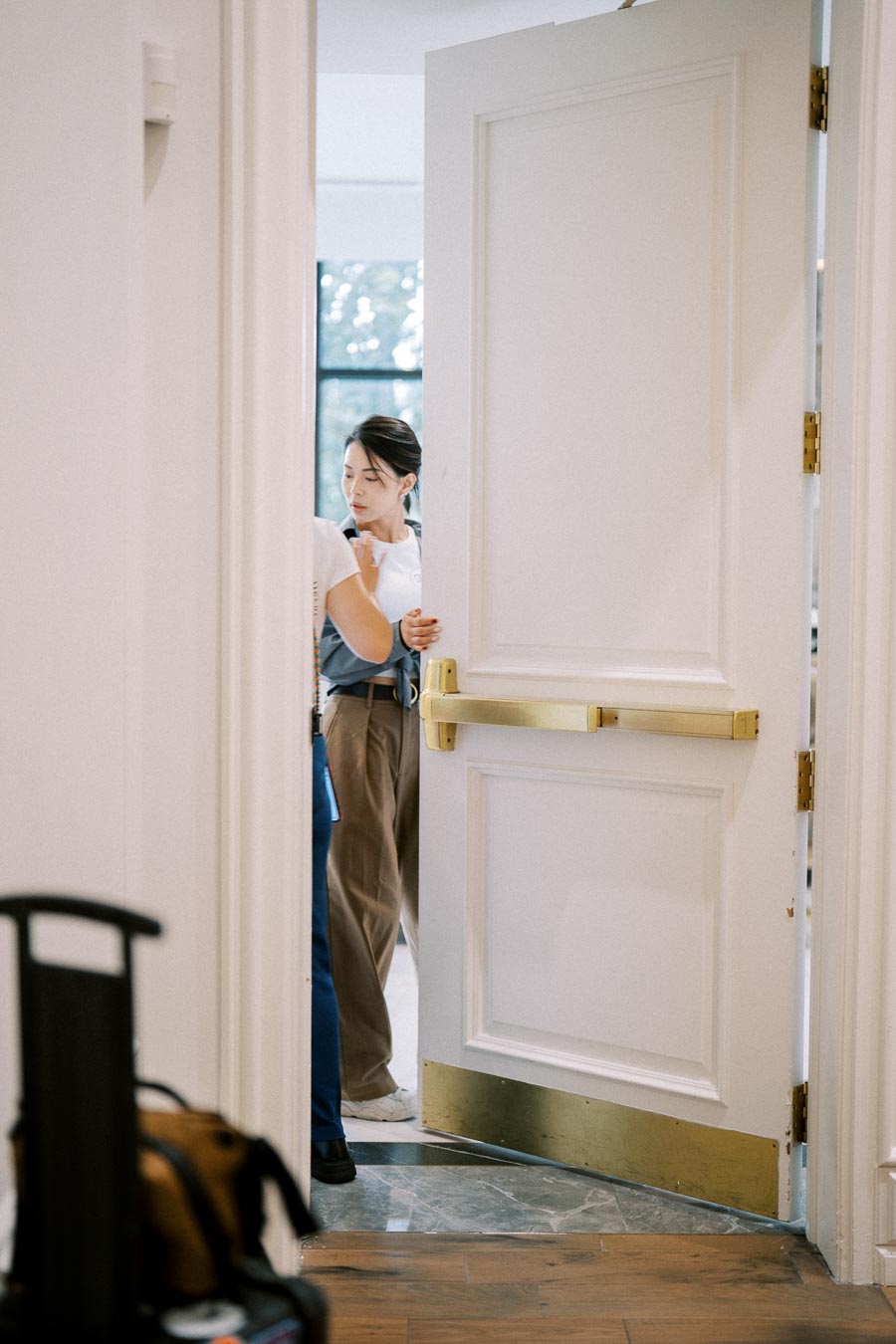 Person entering through a partially open door in a bright, modern interior with natural light coming through a window in the background.